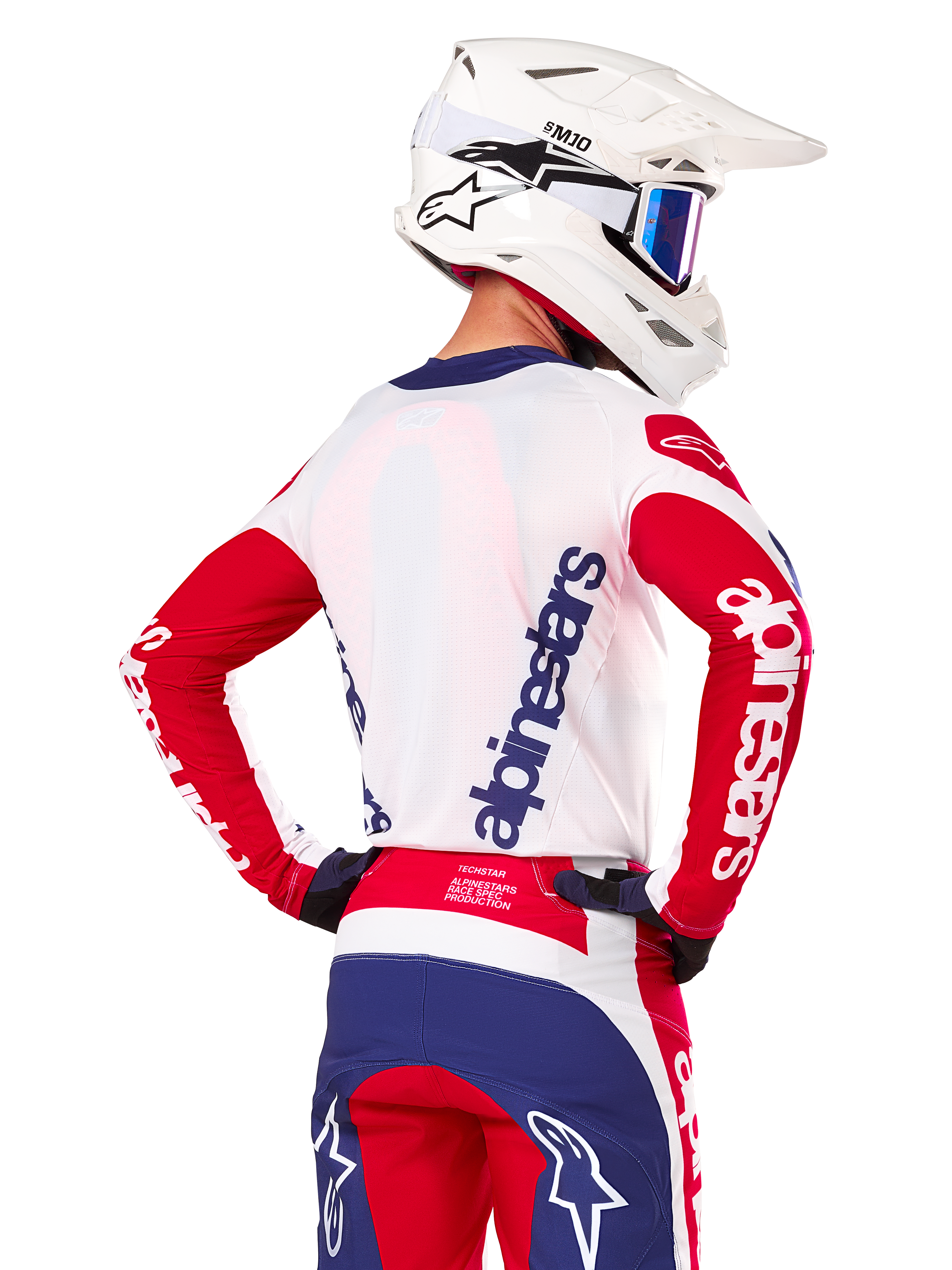 Jersey Alpinestars Techstar Fortitude Edición Limitada, jersey de motocross, rojo brillante, blanco y azul, vista trasera de un piloto con casco blanco y equipamiento a juego, con logotipos llamativos de Alpinestars en la espalda y las mangas, diseñado para carreras off-road de alto rendimiento.