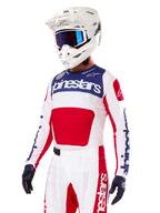 Piloto de motocross con el Jersey Alpinestars Techstar Fortitude Edición Limitada, en rojo brillante, blanco y azul, con casco blanco, gafas azules y pantalones a juego, equipación de competición de alto rendimiento para off-road
