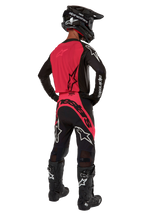 Piloto de motocross desde atrás con el Jersey Supertech Ward 2024 en Negro Red Berry, con un vibrante panel trasero rojo y mangas negras con la marca Alpinestars en blanco. Combinado con pantalones de motocross a juego, botas Tech 10 negras y un casco de fibra de carbono negro para un conjunto de equipamiento de carreras profesional coordinado.