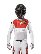 Piloto de motocross con el jersey Alpinestars Supertech Ward 2024 en White Hot Orange, jersey de malla elástica blanca con un gráfico rojo prominente en el pecho y el logo, casco negro con gafas oscuras, guantes blancos, equipamiento de nivel profesional para rendimiento off-road