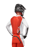 Piloto de motocross con el Jersey Alpinestars Supertech Ward 2024 en White Hot Orange, vista trasera que muestra el panel posterior naranja y mangas blancas con la marca en negro, combinado con pantalones a juego y casco negro, equipamiento profesional para carreras todoterreno