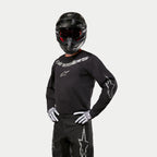 Motocross rider wearing Alpinestars 2024 Fluid Graphite Jersey, Black and Silver, paired with black motocross pants, black helmet with red-tinted goggles, and white and black gloves, featuring silver brand logos on the chest and sleeves.