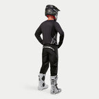 Motocross rider wearing the Alpinestars 2024 Fluid Graphite Jersey in Black Silver from a rear view, long-sleeve athletic fit jersey paired with black motocross pants, black helmet, and white and black boots, complete professional gear for off-road racing.