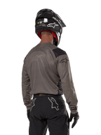 Motocross rider from the back wearing the Alpinestars Techdura Jersey in Falcon Brown, paired with black off-road pants and a black helmet, featuring a modern graphic design and elongated rear for enduro riding
