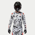 Alpinestars 2024 Techstar Rantera Jersey, jersey de motocross, gris camuflaje con patrones irregulares negros y detalles en rojo, usado por un piloto con pantalones a juego, casco negro y guantes, equipo de carreras todoterreno.