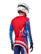 Piloto de motocross visto desde atrás luciendo el Jersey Alpinestars Techstar Pneuma 2024 en azul, rojo marte y blanco, con espalda roja y logo de estrella blanco, mangas azules y moradas con marca en blanco, combinado con pantalones a juego y casco blanco para carreras off-road.