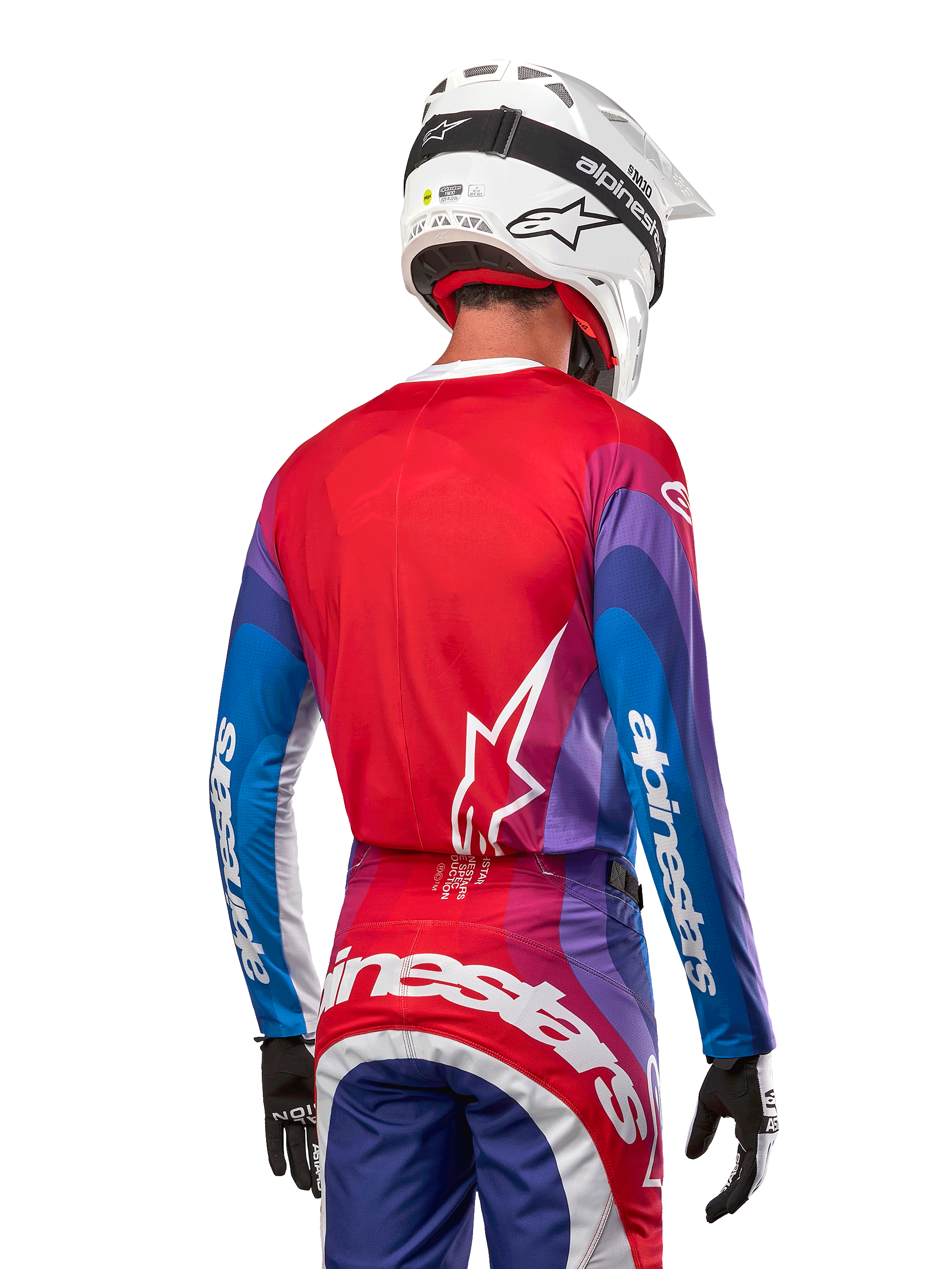 Piloto de motocross visto desde atrás luciendo el Jersey Alpinestars Techstar Pneuma 2024 en azul, rojo marte y blanco, con espalda roja y logo de estrella blanco, mangas azules y moradas con marca en blanco, combinado con pantalones a juego y casco blanco para carreras off-road.