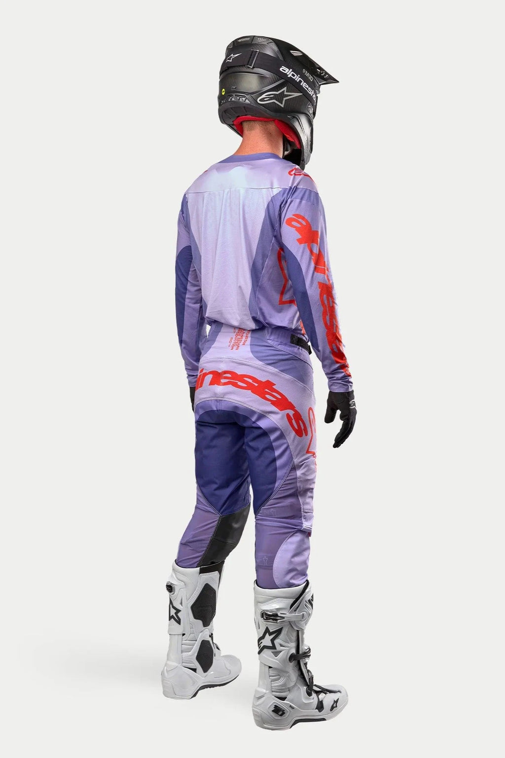 Piloto de motocross con equipamiento de competición completo visto desde atrás, Jersey Alpinestars Techstar Ocuri 2024 púrpura con logotipos en naranja, pantalones púrpuras a juego con detalles naranjas, casco negro, botas blancas y negras, guantes negros, kit profesional de carreras off-road