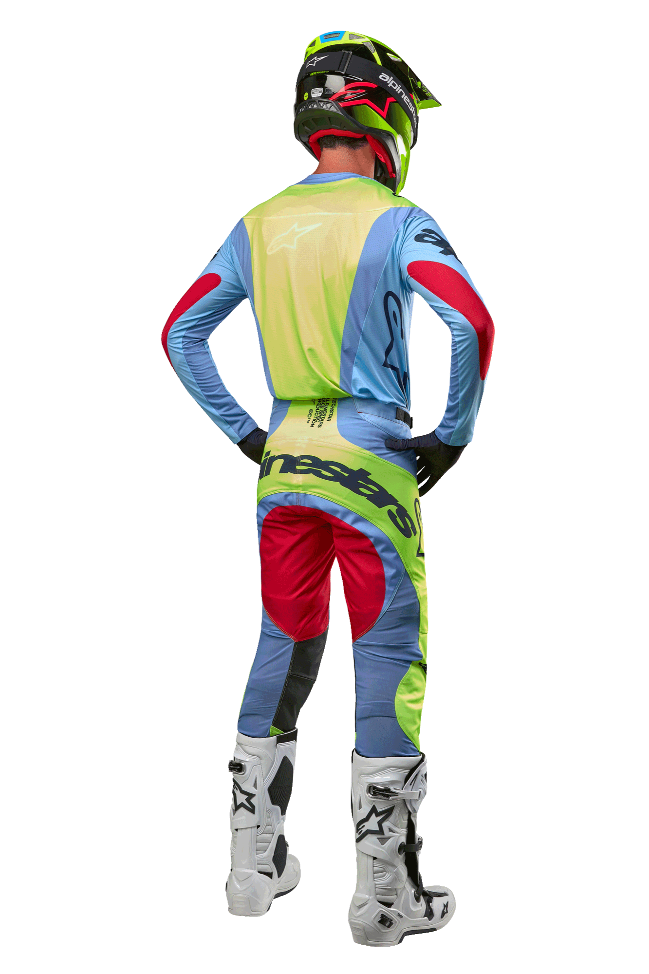 Piloto de motocross visto desde atrás con el jersey Alpinestars Techstar Ocuri 2024, azul claro con detalles en amarillo flúor y rojo baya, pantalones de carreras a juego, botas blancas y casco neón, equipamiento completo de off-road para competición profesional
