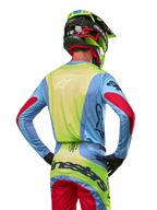 Piloto de motocross desde atrás vistiendo el Jersey Alpinestars Techstar Ocuri 2024, azul claro con espalda en amarillo flúor y detalles rojos en las mangas, pantalones de motocross a juego y casco amarillo neón, conjunto coordinado para carreras fuera de carretera