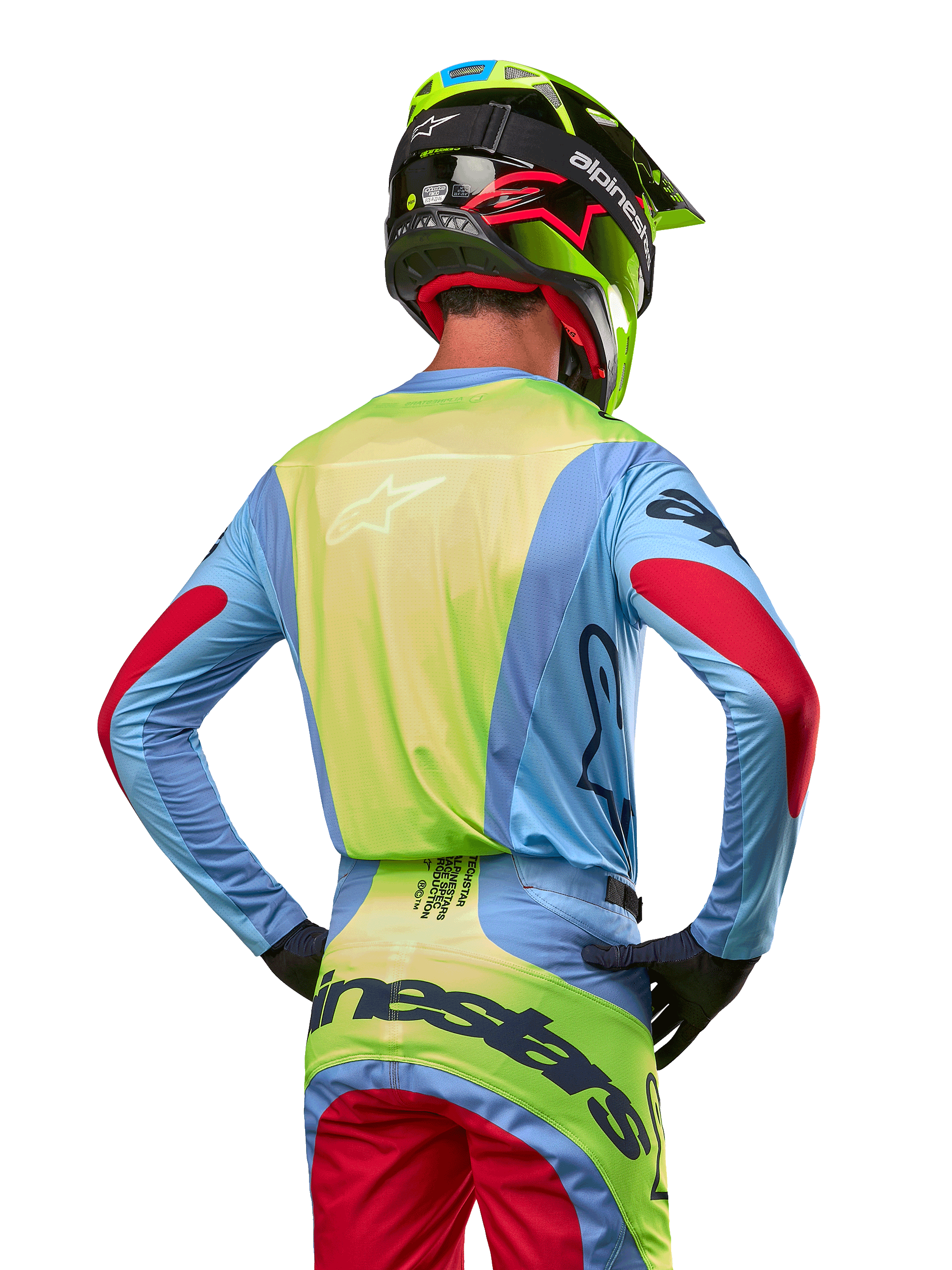 Piloto de motocross desde atrás vistiendo el Jersey Alpinestars Techstar Ocuri 2024, azul claro con espalda en amarillo flúor y detalles rojos en las mangas, pantalones de motocross a juego y casco amarillo neón, conjunto coordinado para carreras fuera de carretera