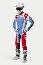 Piloto de motocross con equipamiento completo de carreras, incluyendo el jersey Alpinestars 2024 Techstar Ocuri en azul claro, rojo Marte y blanco, pantalones a juego, casco blanco con gafas, botas blancas y guantes blancos y negros.