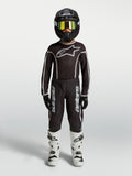 Alpinestars Fluid Apex V2, jersey juvenil y conjunto de motocross, negro y plateado, con botas blancas y casco negro mate.