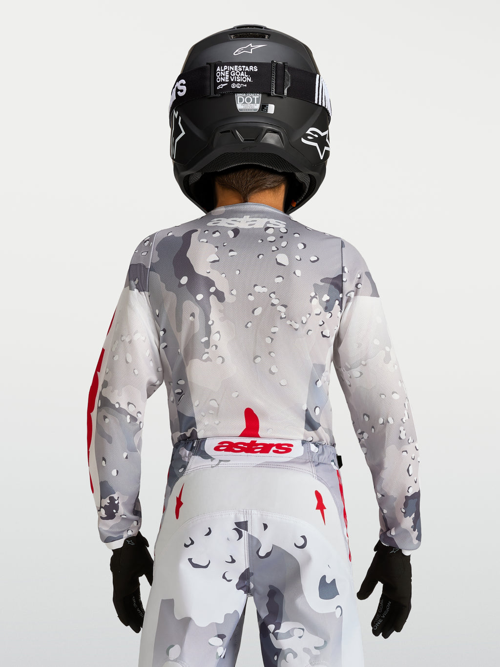 Jersey Fluid Lone juvenil 2027 Alpinestars, gris camuflaje con detalles rojos, vista trasera con casco negro de motocross.