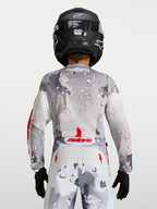 Jersey Fluid Lone juvenil 2027 Alpinestars, gris camuflaje con detalles rojos, vista trasera con casco negro de motocross.