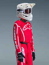 Young motocross rider wearing Alpinestars 2026 Youth Fluid Apex Jersey in Red and White, paired with matching red pants, white helmet, and black gloves, featuring prominent branding and star logo on the chest and sleeves, coordinated outfit for youth off-road racing