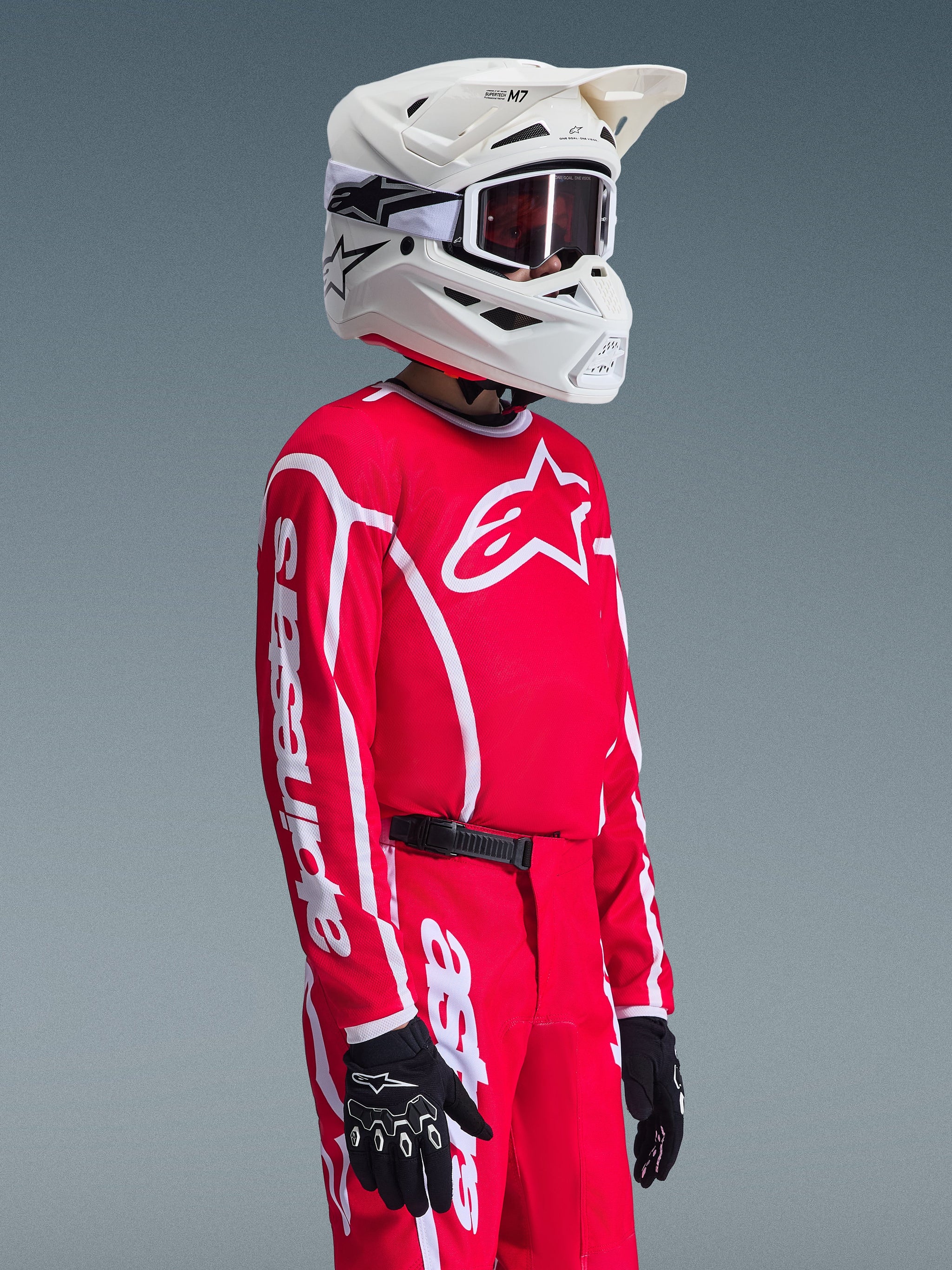 Young motocross rider wearing Alpinestars 2026 Youth Fluid Apex Jersey in Red and White, paired with matching red pants, white helmet, and black gloves, featuring prominent branding and star logo on the chest and sleeves, coordinated outfit for youth off-road racing