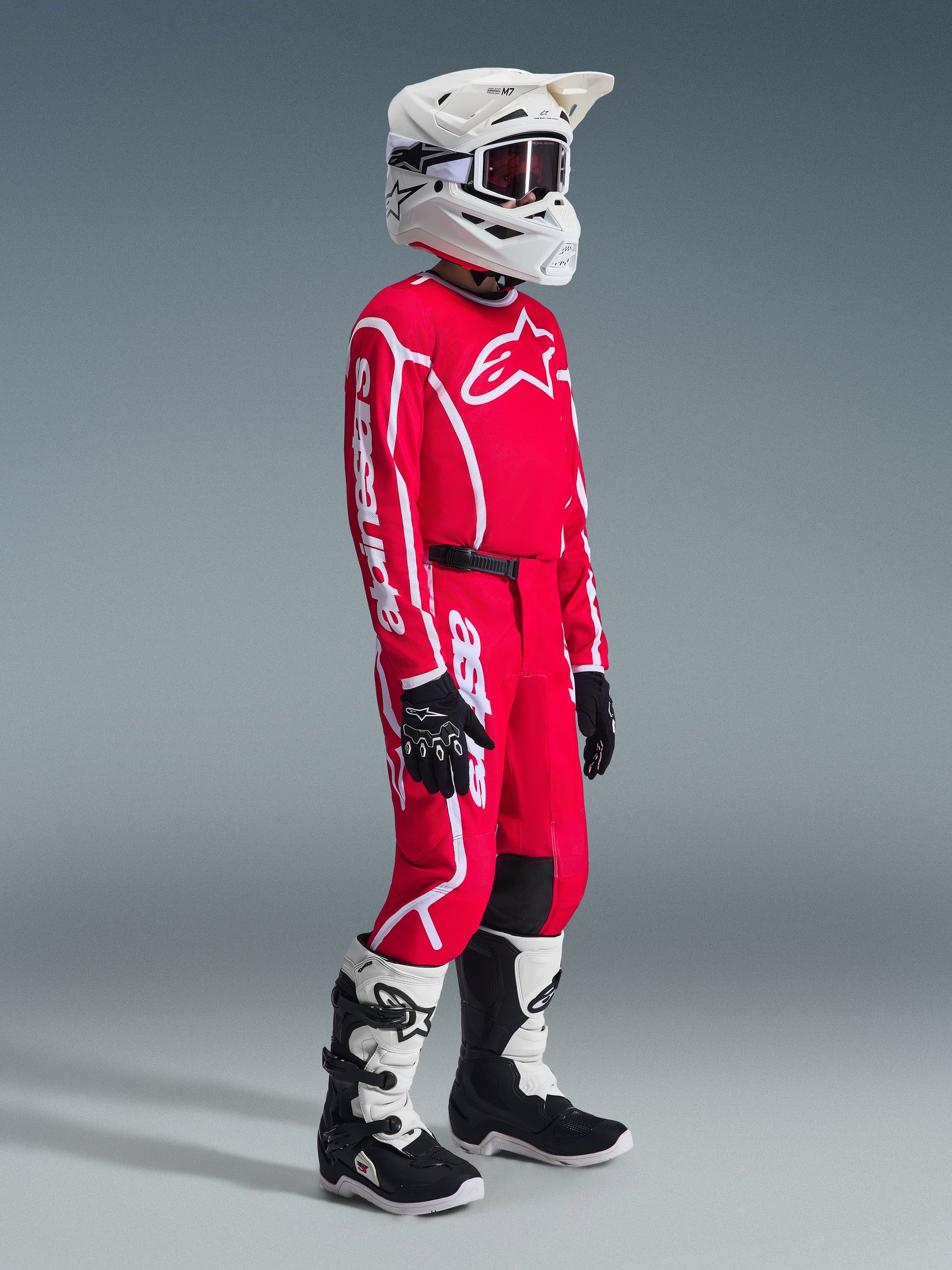 Youth motocross rider wearing complete racing gear, Red 2026 Youth Fluid Apex Jersey with white Alpinestars logos, matching red pants with white graphics and black accents, white helmet with goggles, white and black boots, black and white gloves, coordinated outfit for young off-road racers.