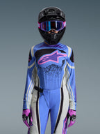 Piloto femenina de motocross con el conjunto Alpinestars 2026 Stella Techstar Nomur, jersey azul con logotipo de estrella rosa y morado, pantalones azules con paneles laterales blancos y negros, casco negro con gafas moradas y azules, y guantes rosas y negros, equipamiento completo de competición off-road para mujer