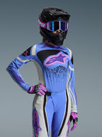 Piloto femenina de motocross con el conjunto Alpinestars 2026 Stella Techstar Nomur, jersey azul claro con detalles en rosa y morado, pantalones azules y blancos a juego, casco integral negro con gafas moradas y guantes rosas y negros, equipamiento profesional de carreras off-road para mujer.