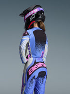 Vista trasera de una piloto de motocross con el conjunto Alpinestars Stella Techstar Nomur 2026, color azul claro con detalles rosa y negro, jersey y pantalones a juego, casco negro con gafas rosas, ajuste ergonómico femenino para competición todoterreno