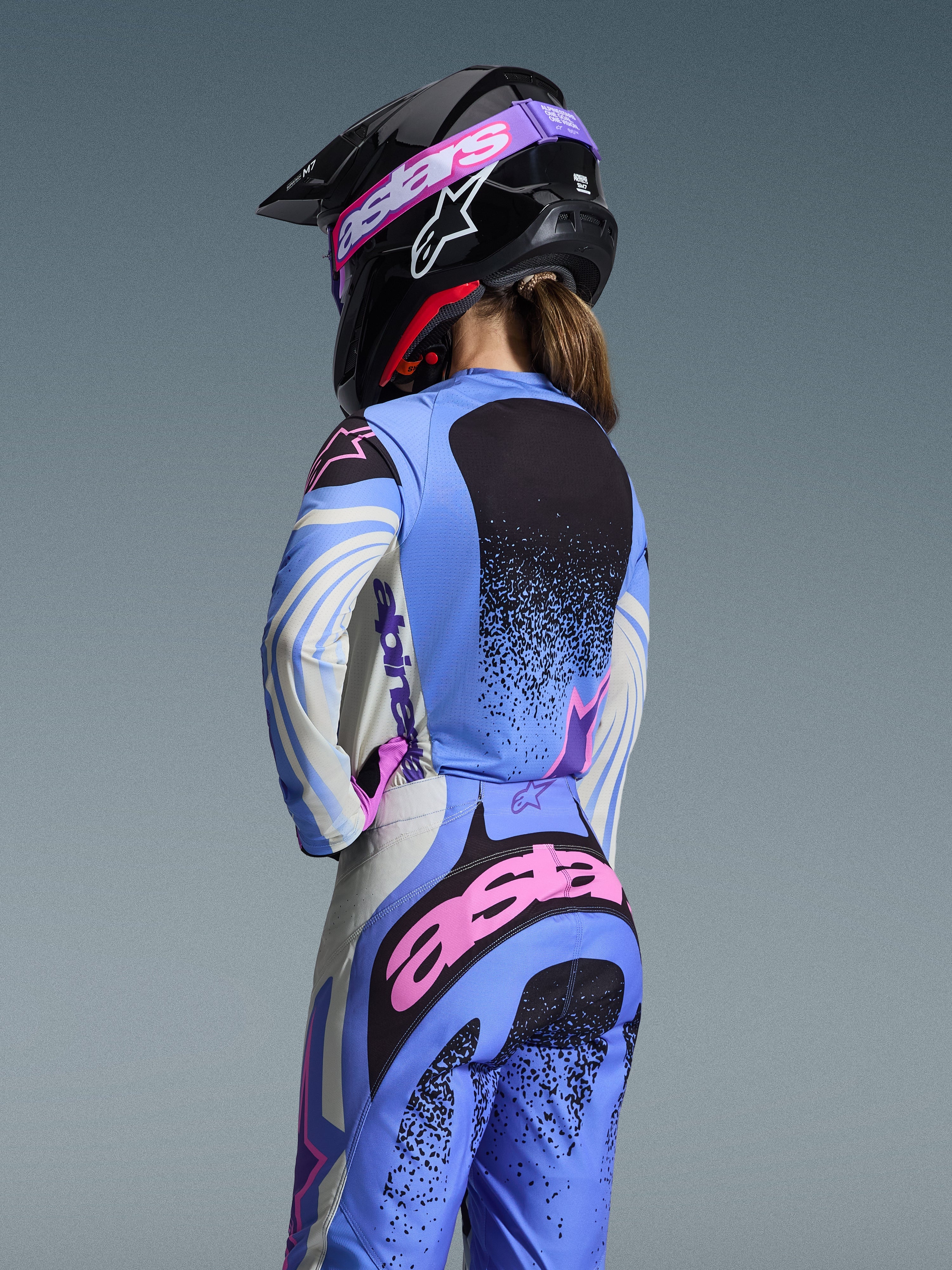Vista trasera de una piloto de motocross con el conjunto Alpinestars Stella Techstar Nomur 2026, color azul claro con detalles rosa y negro, jersey y pantalones a juego, casco negro con gafas rosas, ajuste ergonómico femenino para competición todoterreno