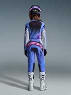 Vista posterior de una piloto de motocross con el conjunto Alpinestars 2026 Stella Techstar Nomur, jersey y pantalones azules con detalles rosas y blancos, logotipo rosa llamativo en la parte trasera de los pantalones, botas de motocross blancas y casco coordinado en negro y rosa.