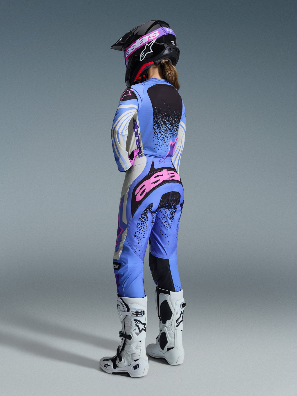 Alpinestars 2026 Stella Techstar Nomur Gear Set, vista trasera de una piloto de motocross con jersey y pantalones en azul claro, blanco y rosa con detalles moteados en negro, combinado con un casco negro y botas blancas, equipamiento profesional de carreras para mujer.
