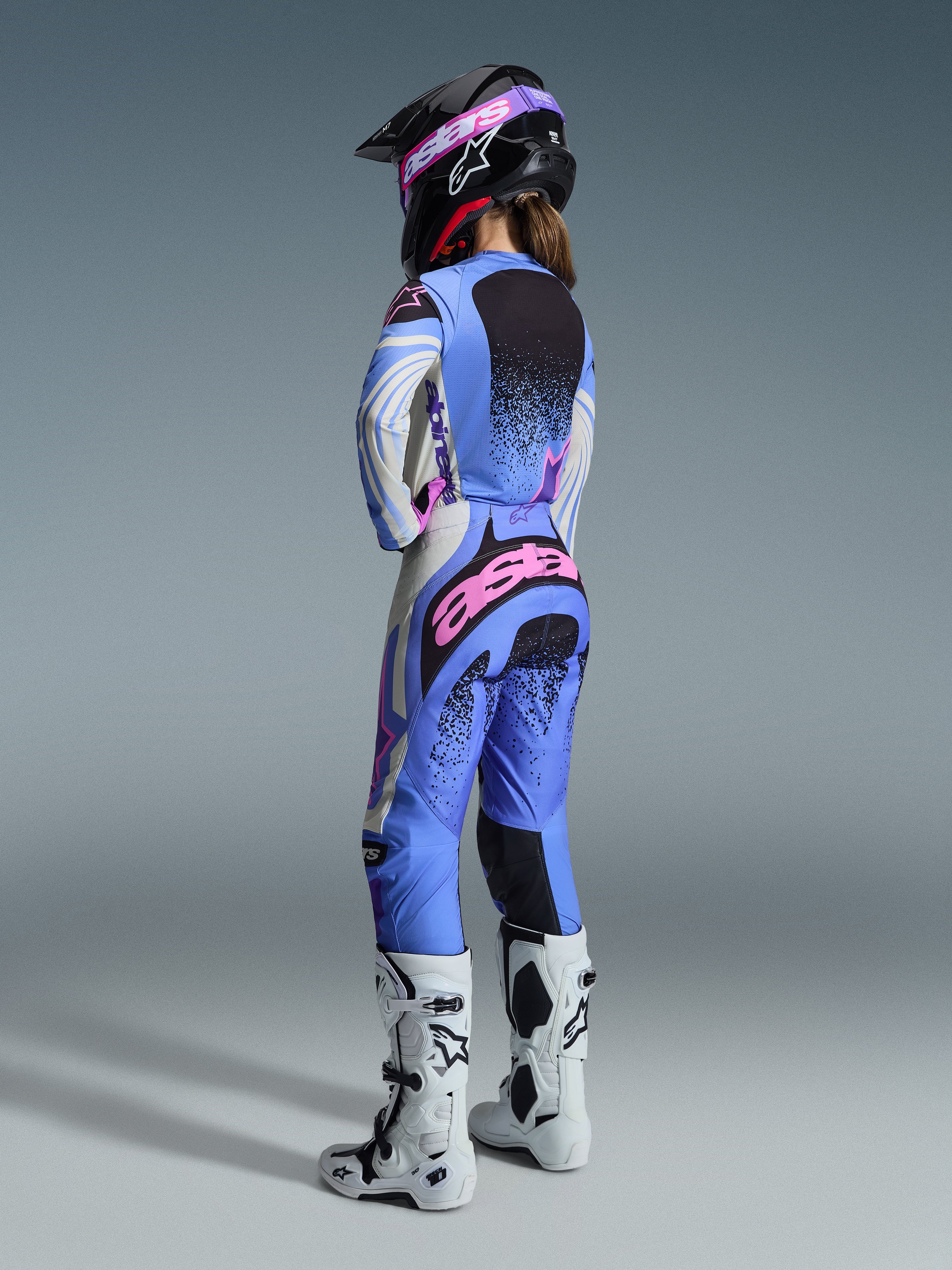 Alpinestars 2026 Stella Techstar Nomur Gear Set, vista trasera de una piloto de motocross con jersey y pantalones en azul claro, blanco y rosa con detalles moteados en negro, combinado con un casco negro y botas blancas, equipamiento profesional de carreras para mujer.