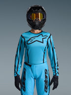Piloto de motocross con el conjunto Stella Fluid Apex 2026 de Alpinestars para mujer, jersey azul claro con detalles y logotipo en negro, pantalones a juego, casco negro con gafas doradas iridiscentes y guantes negros, equipamiento de carreras coordinado.