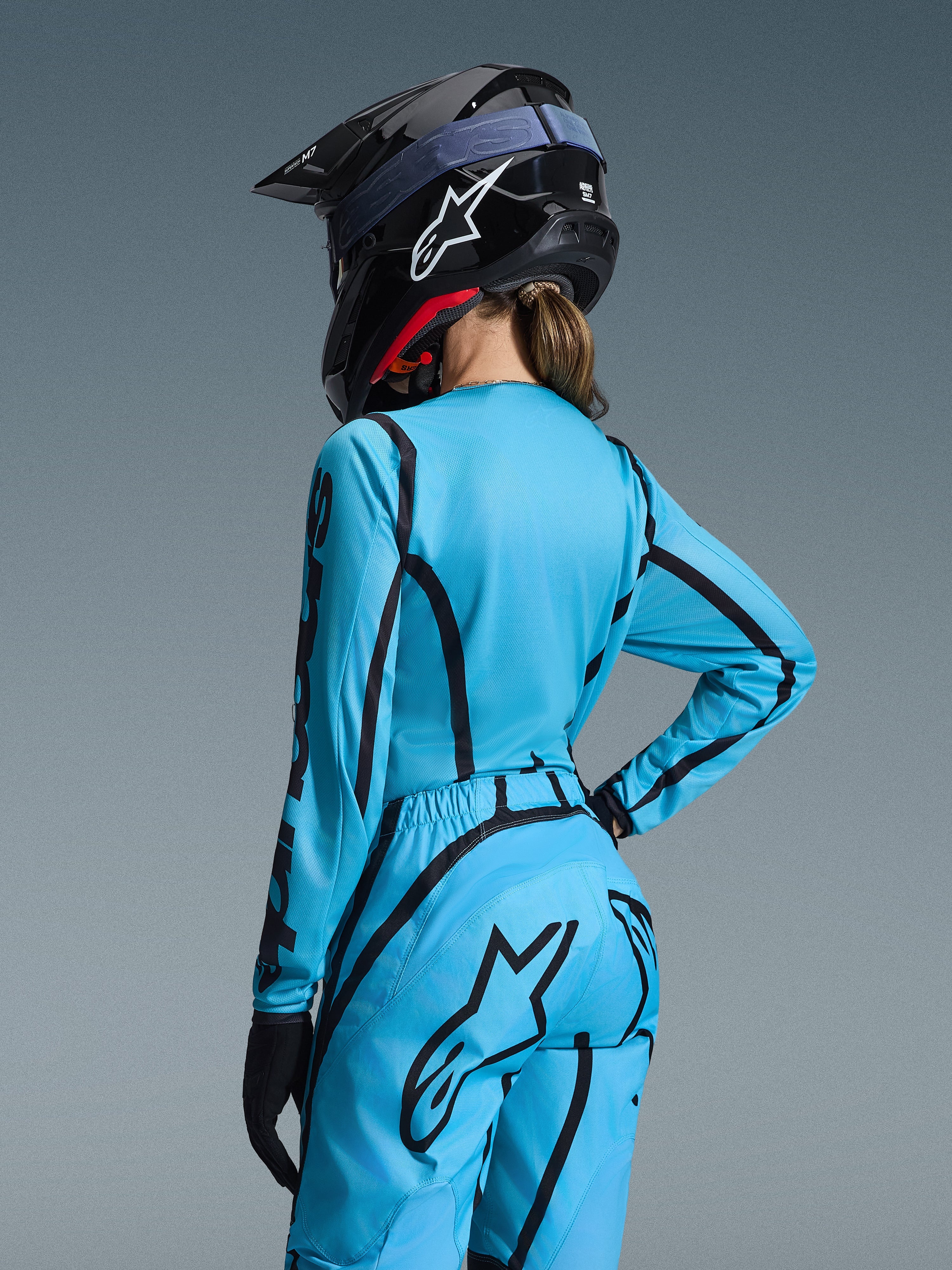 Conjunto de equipamiento Alpinestars Stella Fluid Apex 2026, azul claro y negro, vista posterior de una pilota con jersey de motocross y pantalones a juego, casco Alpinestars negro con logotipo blanco, indumentaria técnica de carreras off-road para mujer.