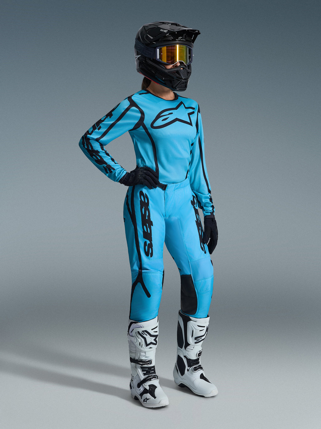 Alpinestars 2026 Woman Stella Fluid Apex Gear Set en Tropical Black, con jersey azul claro y pantalones a juego con detalles en negro, usado por una pilota con casco negro, gafas iridiscentes y botas blancas, equipo profesional de motocross.