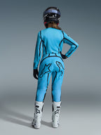 Piloto de motocross vista desde atrás, con el conjunto de equipo Stella Fluid Apex 2026 de Alpinestars para mujer en azul brillante con detalles de líneas gráficas en negro, con un gran logotipo de estrella negra en el pantalón, combinado con botas blancas y negras y un casco negro.