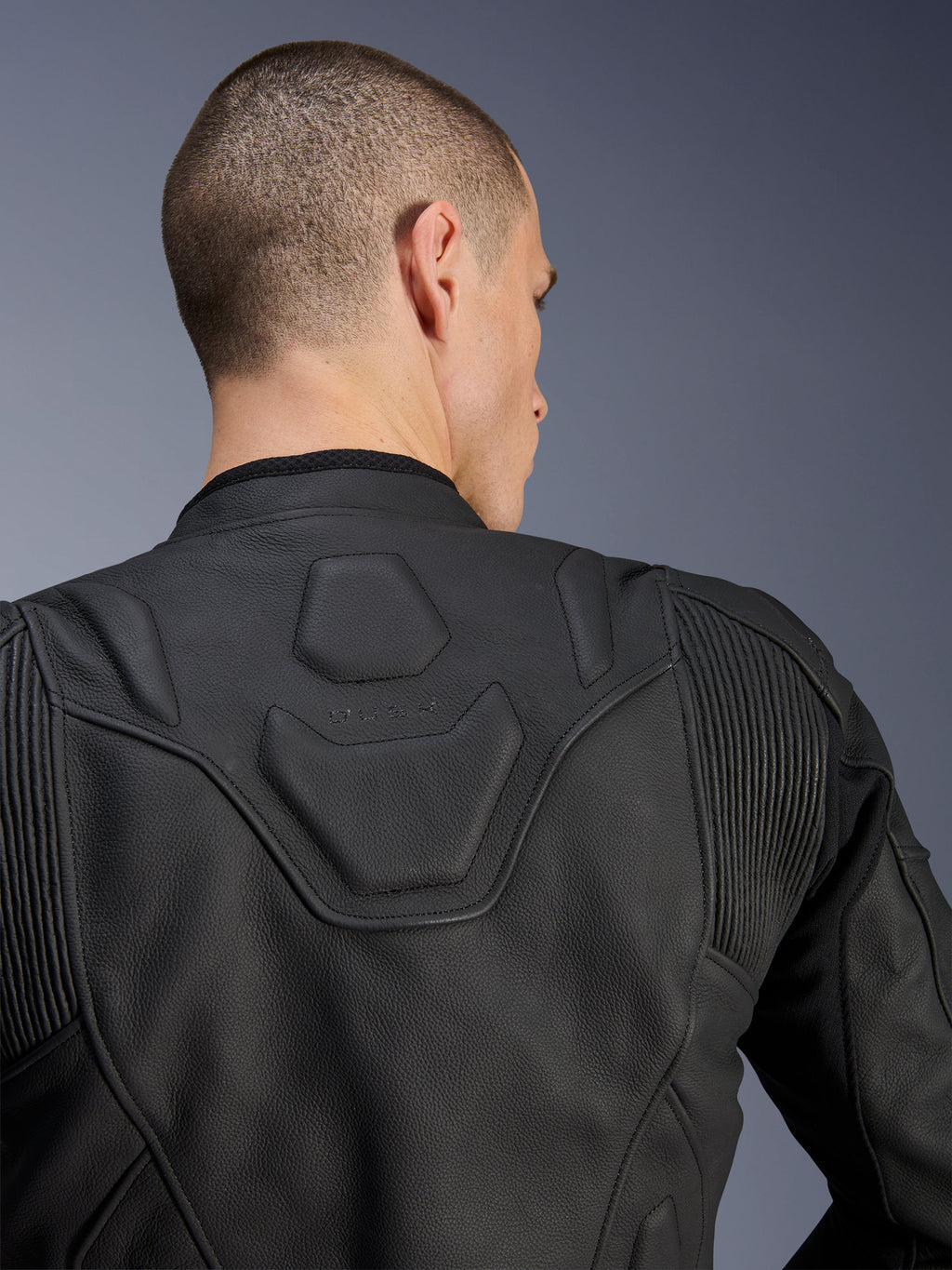 Chaqueta de cuero Alpinestars Dusk, negro, primer plano de la parte superior de la espalda y los hombros con el logotipo DUSK grabado en relieve en un panel acolchado, inserciones elásticas de cuero en acordeón, confeccionada en cuero bovino de primera calidad