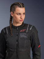 Chaqueta Alpinestars Stella ST-2 Air, chaqueta de moto para mujer en color negro, con paneles de malla para el flujo de aire y marca 'Alpinestars Exploration Equipment' con coordenadas, detalle de logo rojo en el pecho, usada por una modelo con el cabello trenzado.