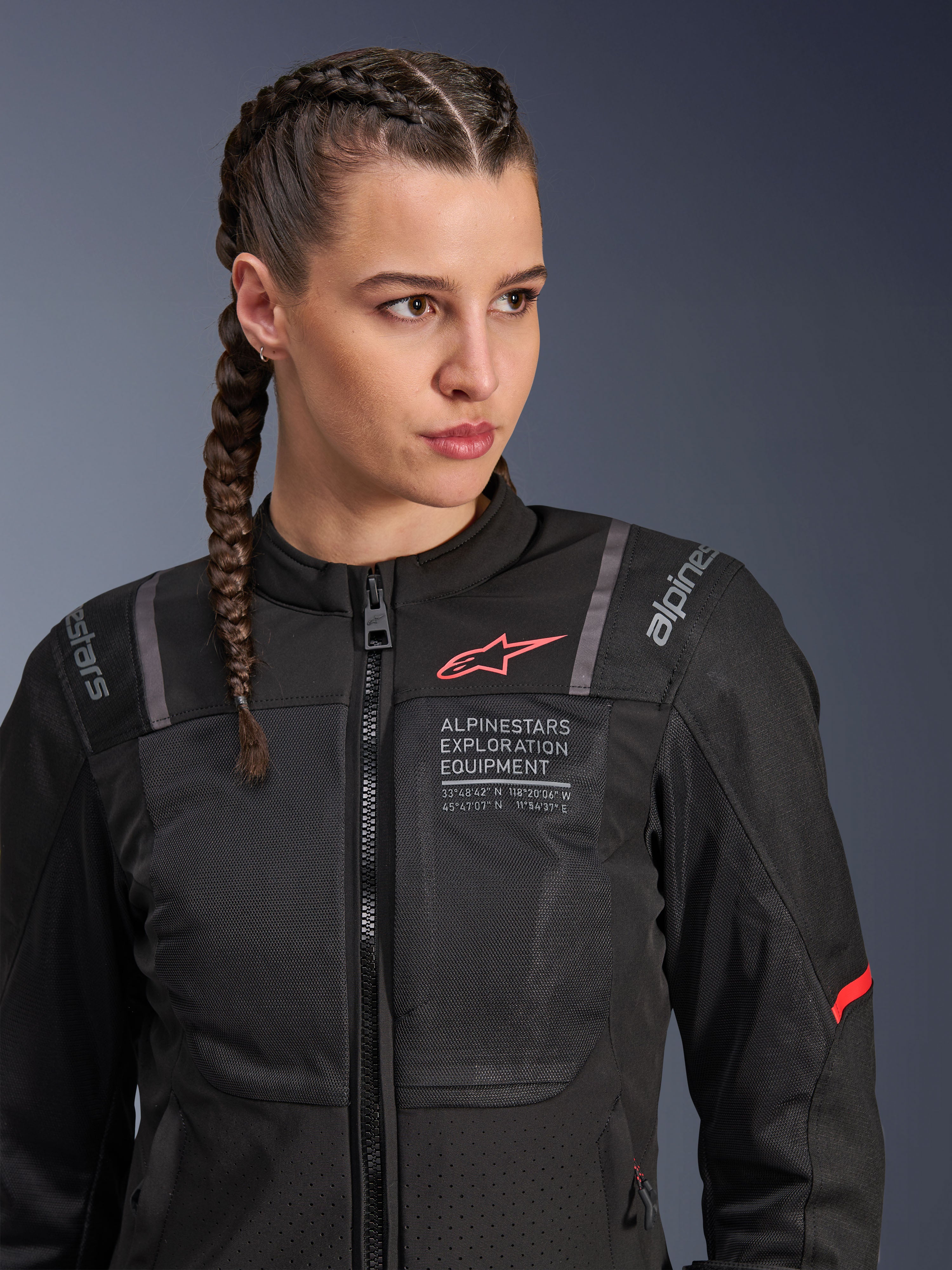 Chaqueta Alpinestars Stella ST-2 Air, chaqueta de moto para mujer en color negro, con paneles de malla para el flujo de aire y marca 'Alpinestars Exploration Equipment' con coordenadas, detalle de logo rojo en el pecho, usada por una modelo con el cabello trenzado.
