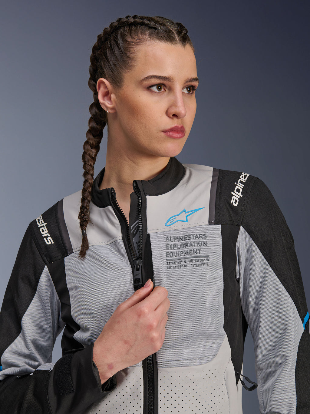 Mujer vistiendo la chaqueta Alpinestars Stella ST-2 Air en gris hielo y negro, retrato de primer plano, con paneles de malla para ventilación, marca 'ALPINESTARS EXPLORATION EQUIPMENT' en el pecho con logotipo azul, chaqueta de moto de aventura moderna