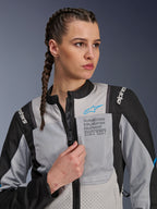 Mujer vistiendo la chaqueta Alpinestars Stella ST-2 Air en gris hielo y negro, retrato de primer plano, con paneles de malla para ventilación, marca 'ALPINESTARS EXPLORATION EQUIPMENT' en el pecho con logotipo azul, chaqueta de moto de aventura moderna
