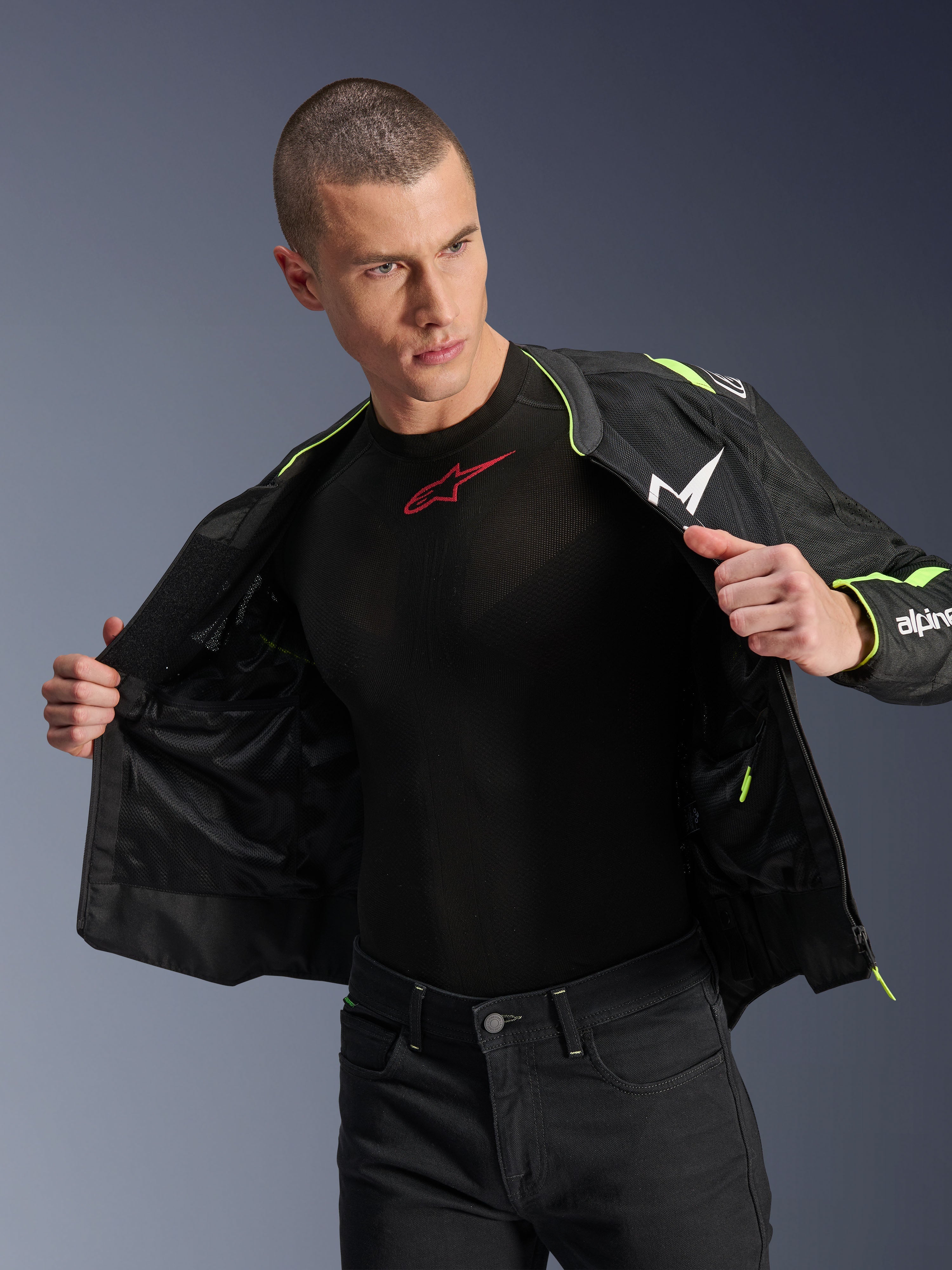 Modelo masculino vistiendo la chaqueta Alpinestars T-Sps Air V2 en negro y amarillo flúor, mostrada abierta para resaltar la construcción interna de malla y los bolsillos, combinada con una prenda técnica Alpinestars negra y pantalones negros, estilo de equipamiento urbano para motociclista