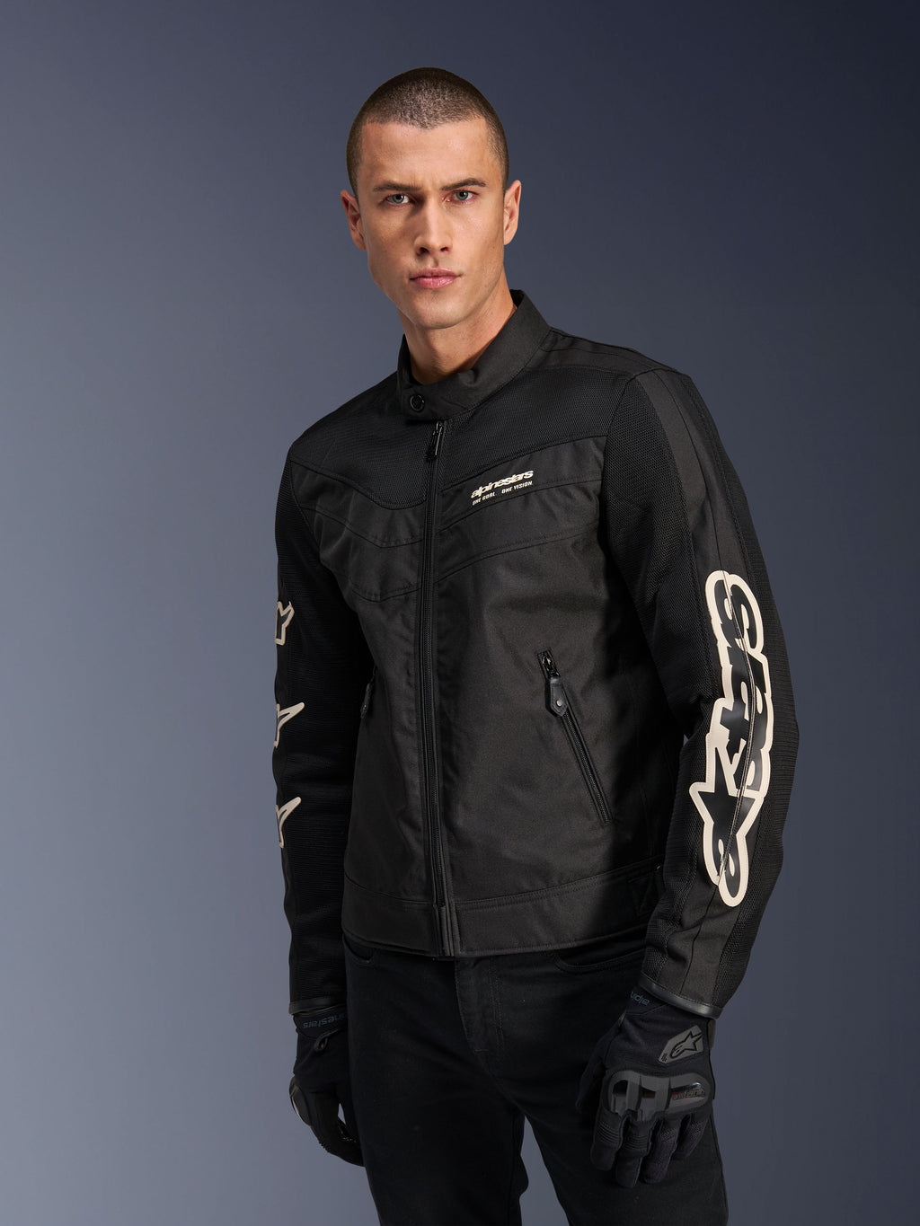 Chaqueta Alpinestars T-Dyno Air en negro, modelo masculino con chaqueta de moto textil de inspiración vintage y paneles de malla para ventilación, logotipo grande de Alpinestars en blanco en las mangas, combinada con guantes de conducción negros, diseño de equipamiento de moto urbano