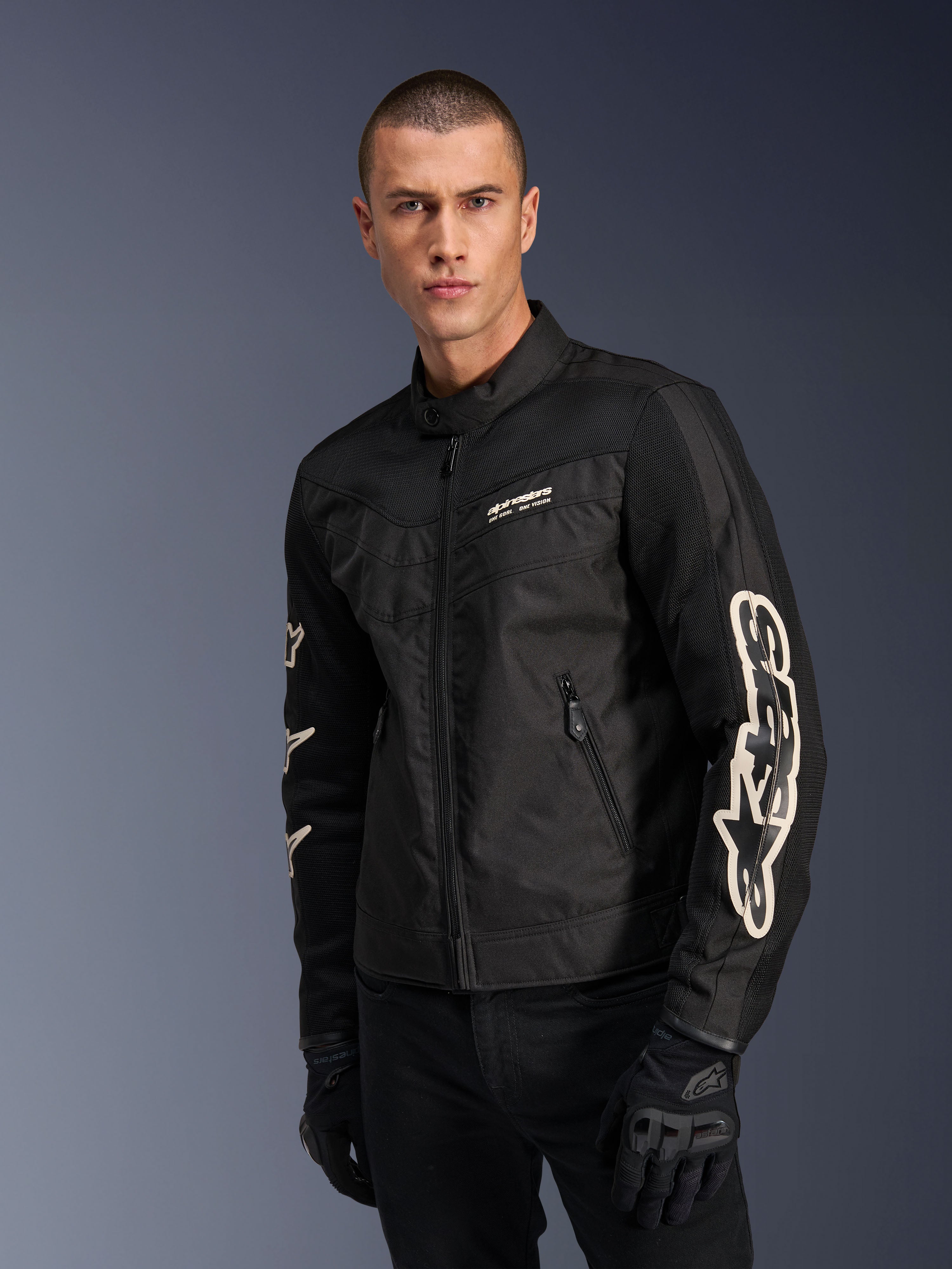 Chaqueta Alpinestars T-Dyno Air en negro, modelo masculino con chaqueta de moto textil de inspiración vintage y paneles de malla para ventilación, logotipo grande de Alpinestars en blanco en las mangas, combinada con guantes de conducción negros, diseño de equipamiento de moto urbano