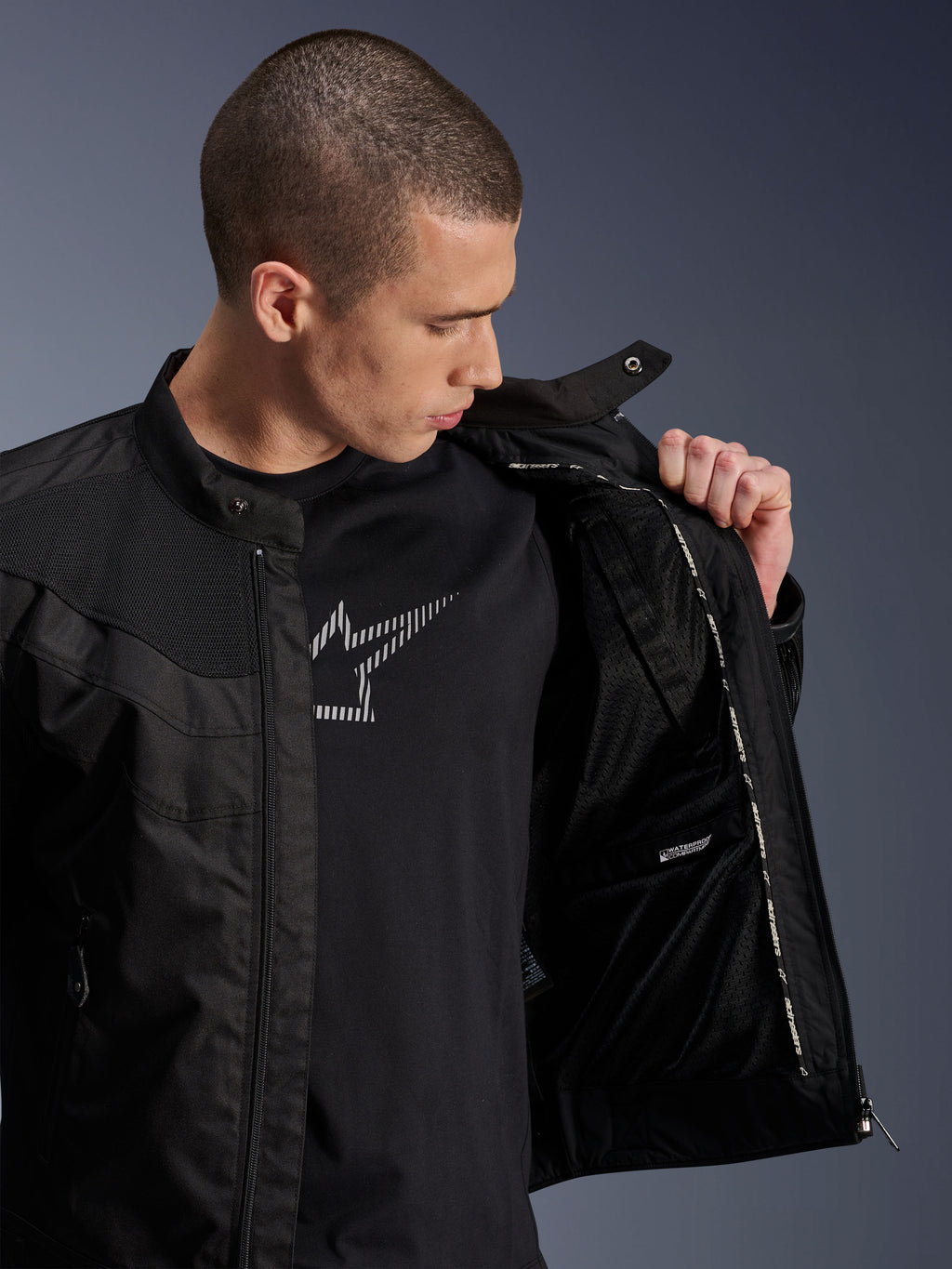 Chaqueta Alpinestars T-Dyno Air, negra, modelo masculino mostrando el forro interno de malla y la etiqueta del compartimento impermeable, sobre una camiseta negra con gráfico, equipamiento moderno para motociclismo urbano