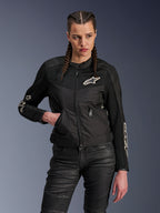 Alpinestars Stella T-Dyno Air Jacket, chaqueta de moto para mujer, negra con logotipos blancos, llevada por una piloto con el pelo trenzado, con paneles de malla 3D transpirable y bolsillos con cremallera, combinada con pantalones de conducción negros texturizados, equipamiento urbano de inspiración vintage