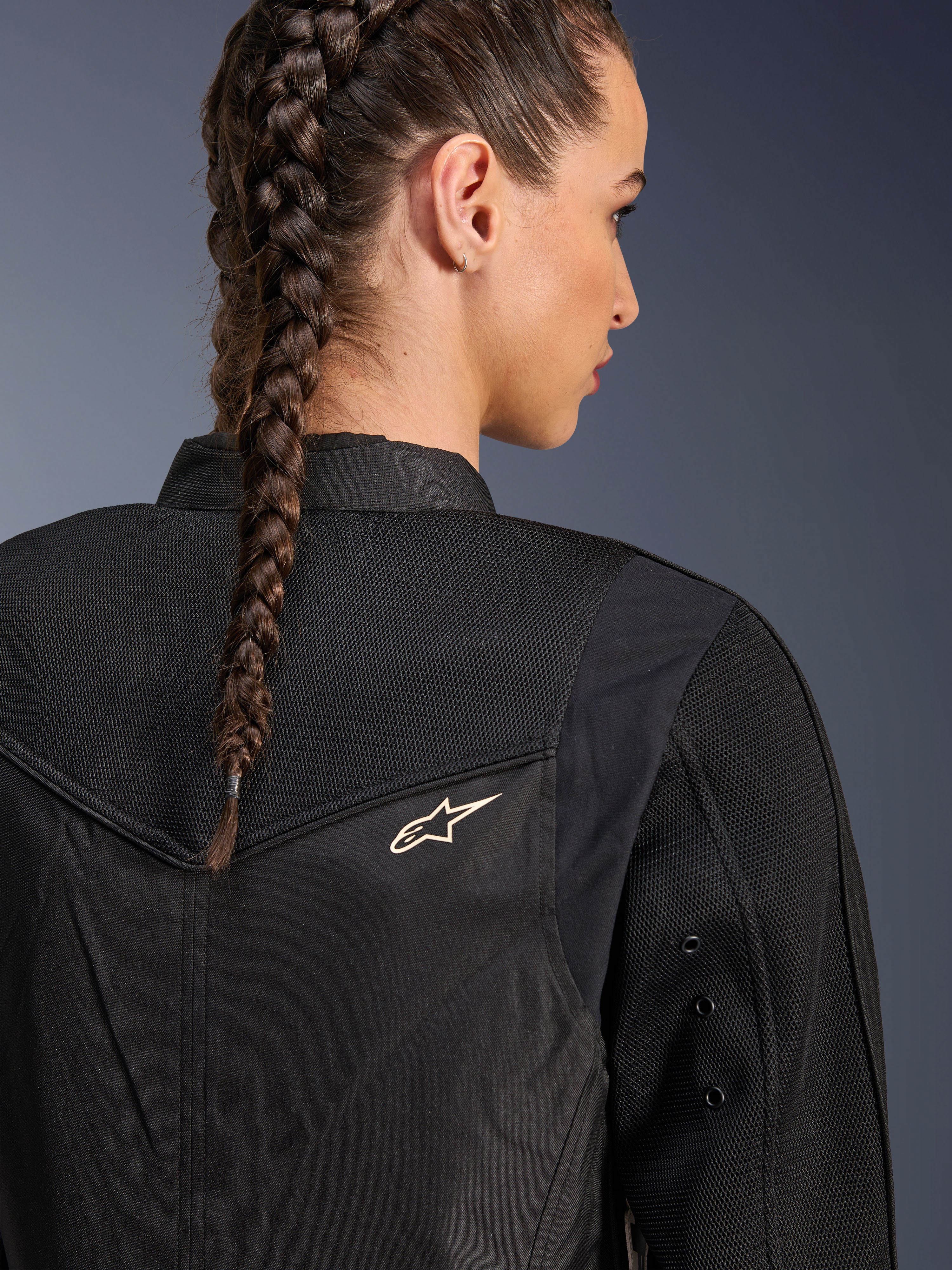 Alpinestars Stella T-Dyno Air Jacket, chaqueta de moto negra para mujer, vista de cerca de la parte posterior que muestra los paneles de malla 3D en el canesú y la parte superior de la espalda para el flujo de aire, con un pequeño logotipo de estrella blanco, lucida por una modelo con el cabello trenzado.