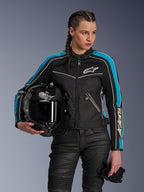 Mujer vistiendo la chaqueta Alpinestars Stella T-Dyno Air en Negro Azul Claro Crudo, sosteniendo un casco de moto de fibra de carbono negro y guantes, con un ajuste entallado, paneles de malla 3D y diseño de inspiración vintage, equipamiento contemporáneo para motociclismo