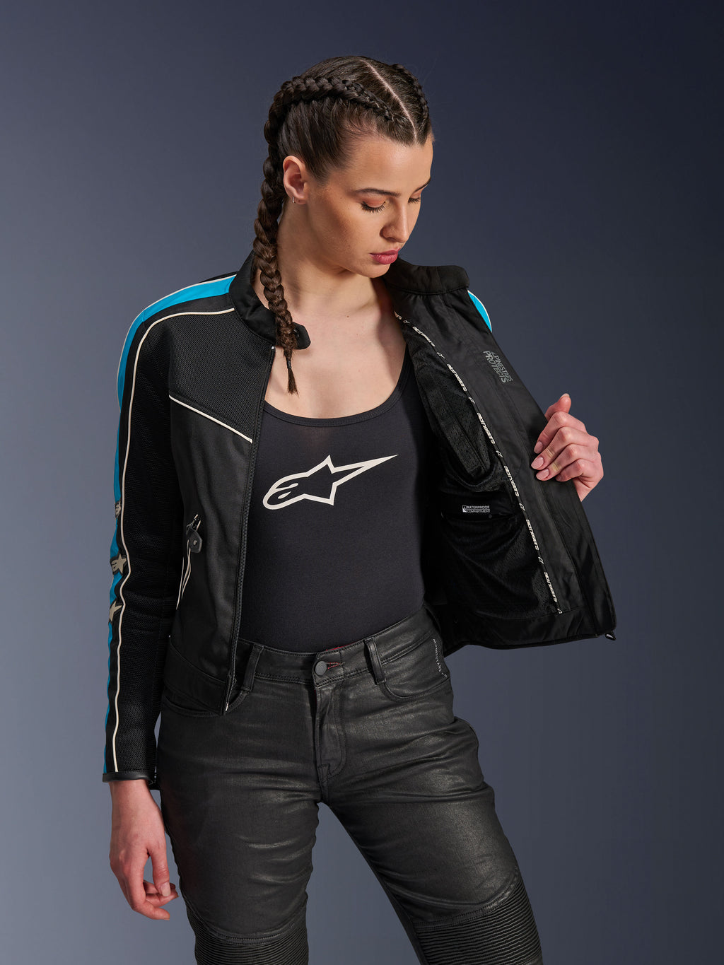 Mujer con la chaqueta Alpinestars Stella T-Dyno Air en Black Light Blue Ecru, abierta para mostrar el forro de malla interior y el bolsillo impermeable, combinada con una camiseta de tirantes negra con logo y pantalones de moto negros, equipamiento urbano de inspiración vintage