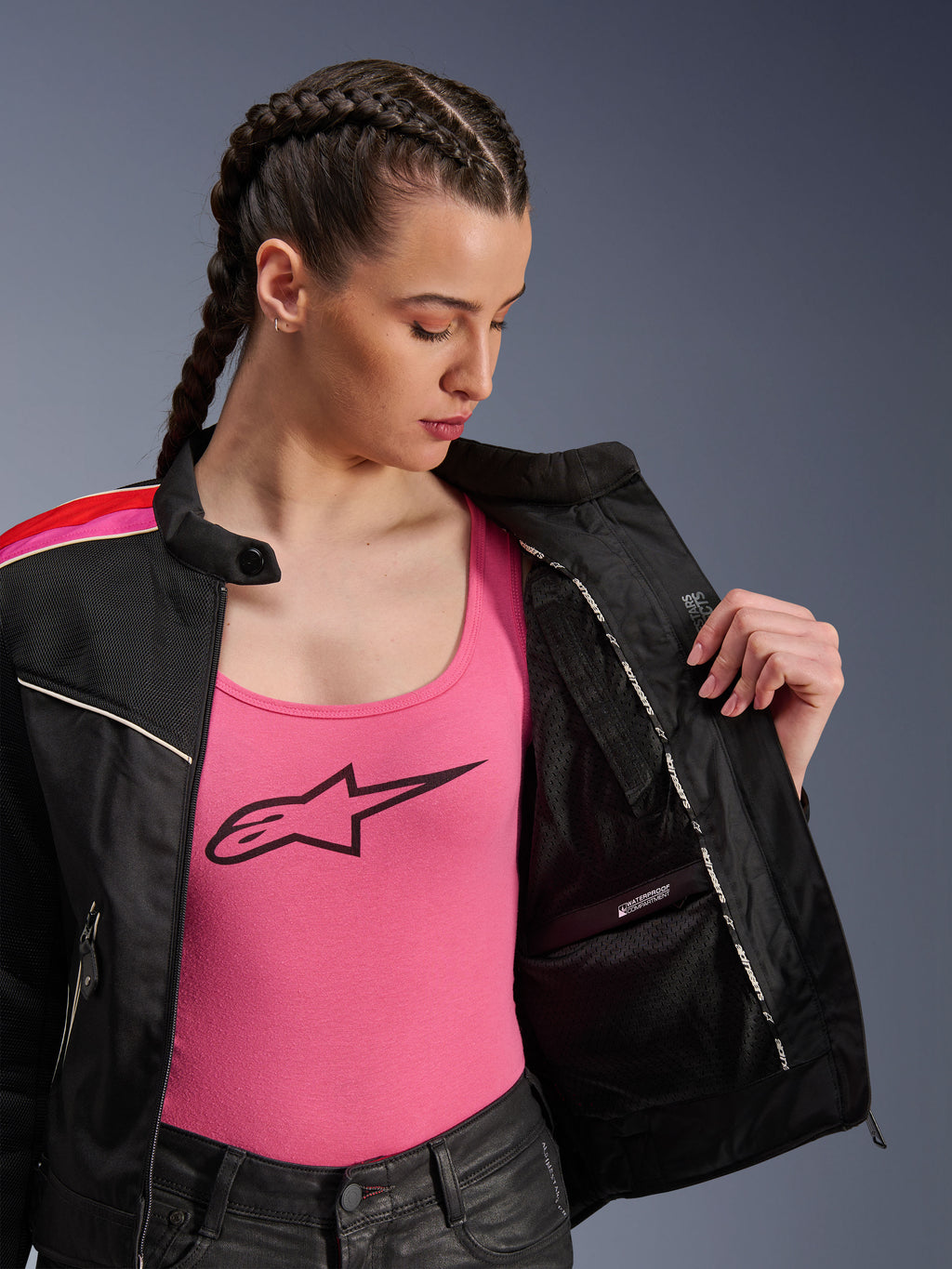 Mujer motociclista vistiendo la chaqueta Alpinestars Stella T-Dyno Air en color Negro Rojo Brillante Fucsia, mostrada parcialmente abierta para revelar el compartimento interno impermeable, usada sobre una camiseta rosa con logo, destacando los paneles de malla transpirable y el ajuste de motociclista entallado.