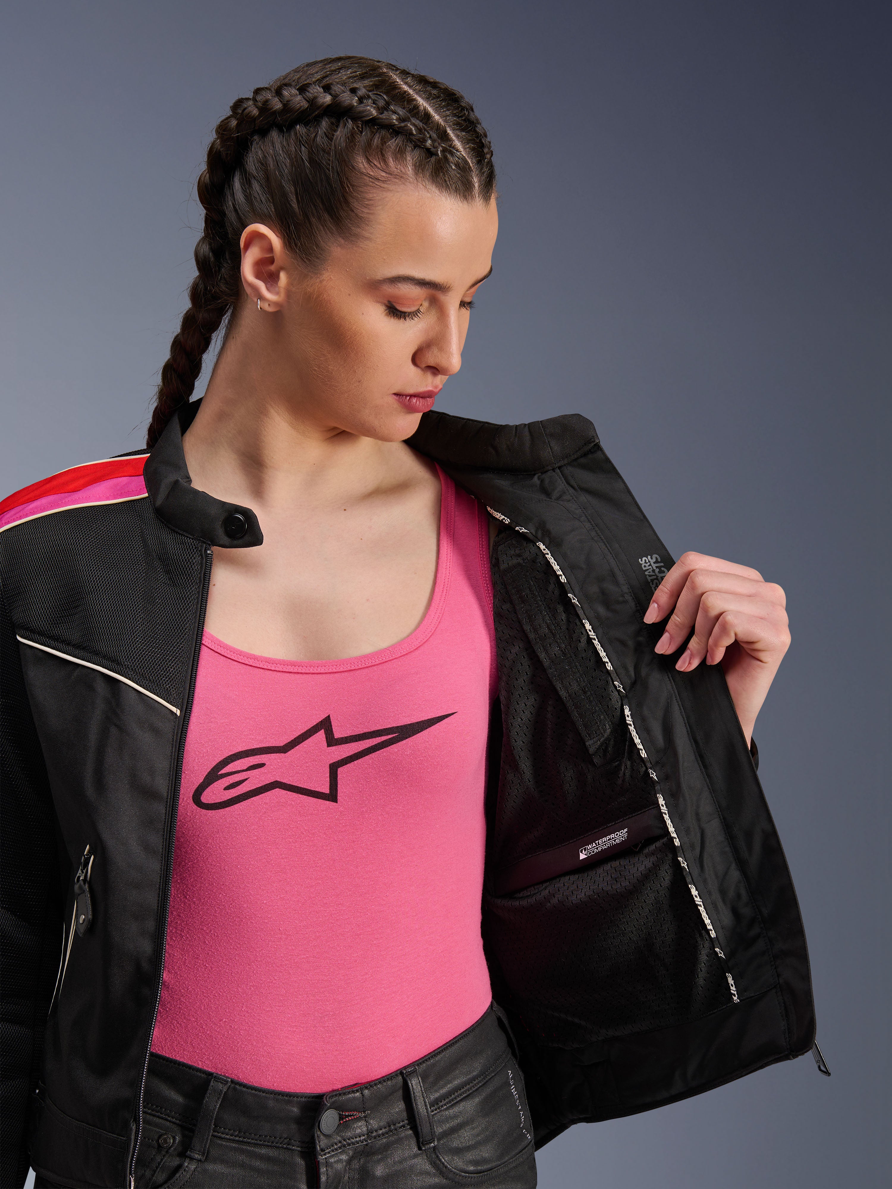Mujer motociclista vistiendo la chaqueta Alpinestars Stella T-Dyno Air en color Negro Rojo Brillante Fucsia, mostrada parcialmente abierta para revelar el compartimento interno impermeable, usada sobre una camiseta rosa con logo, destacando los paneles de malla transpirable y el ajuste de motociclista entallado.