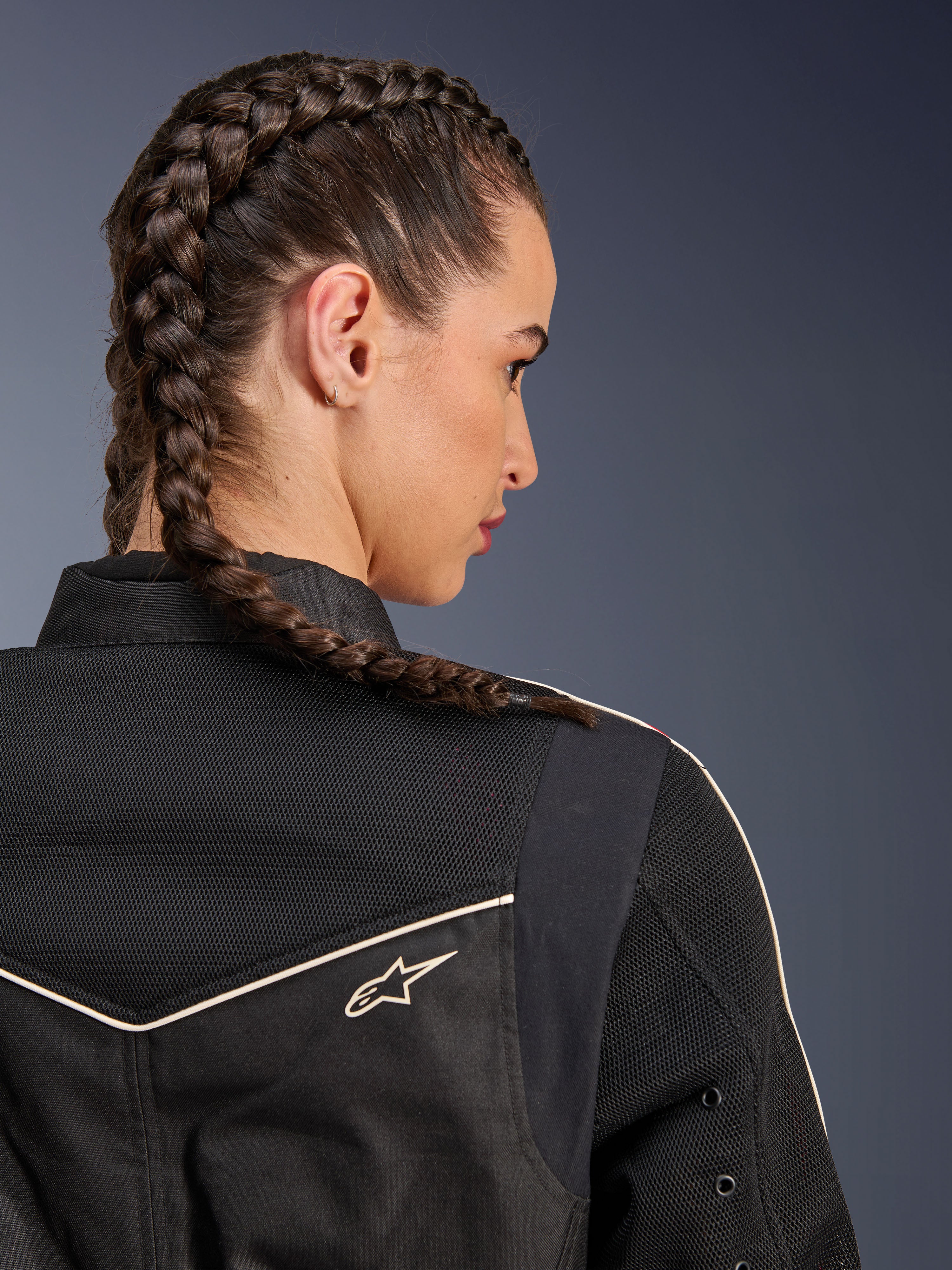 Mujer con la chaqueta Alpinestars Stella T-Dyno Air, primer plano posterior centrado en los hombros y el canesú, negra con paneles de malla 3D transpirable, ribetes blancos y pequeño logotipo de Astars, modelo con el cabello trenzado, ropa urbana para motociclistas