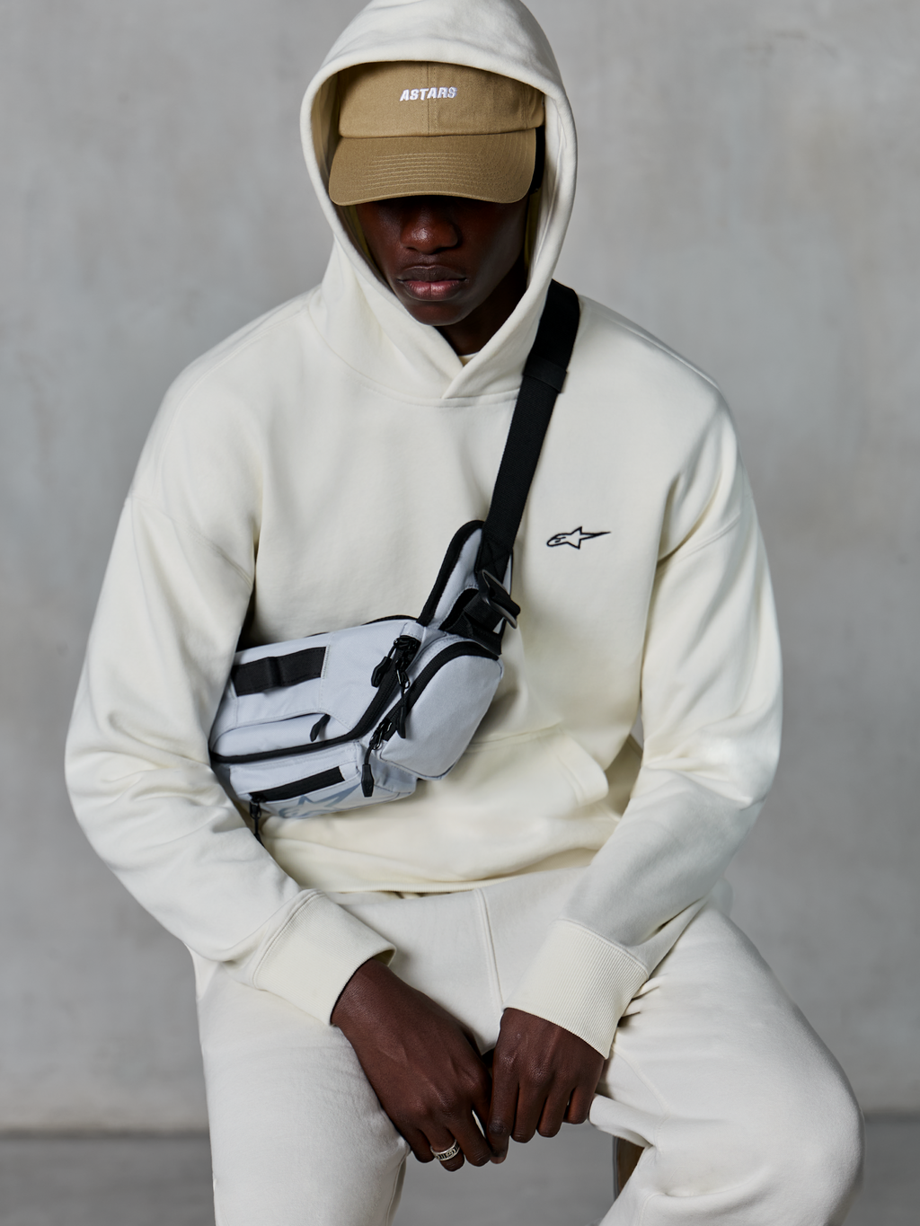 Sudadera con capucha Alpinestars Essentials Ageless, estilo streetwear casual, color blanco roto, pequeño logo negro bordado en el pecho, capucha puesta sobre una gorra color tostado con marca ASTARS, combinada con una bandolera gris claro, ropa lifestyle premium
